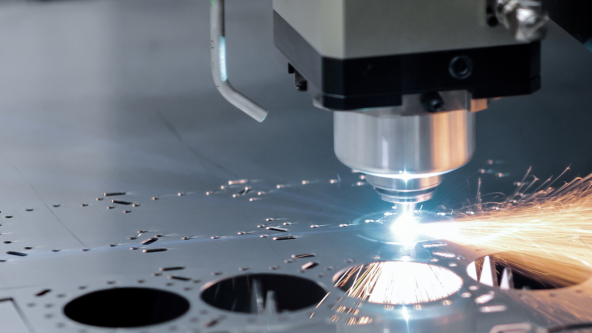 close-up of a CNC laser-cutting head slicing through a flat metal sheet, sending a cascade of bright orange sparks as the beam etches precise circular cutouts on the smooth steel surface